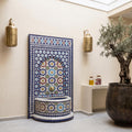 a decorative tile fountain with a mosaic design, flanked by two gold lanterns on the wall. The fountain is situated in a room with a white wall and a wooden floor. A potted plant is also present in the room, adding a touch of greenery to the space.