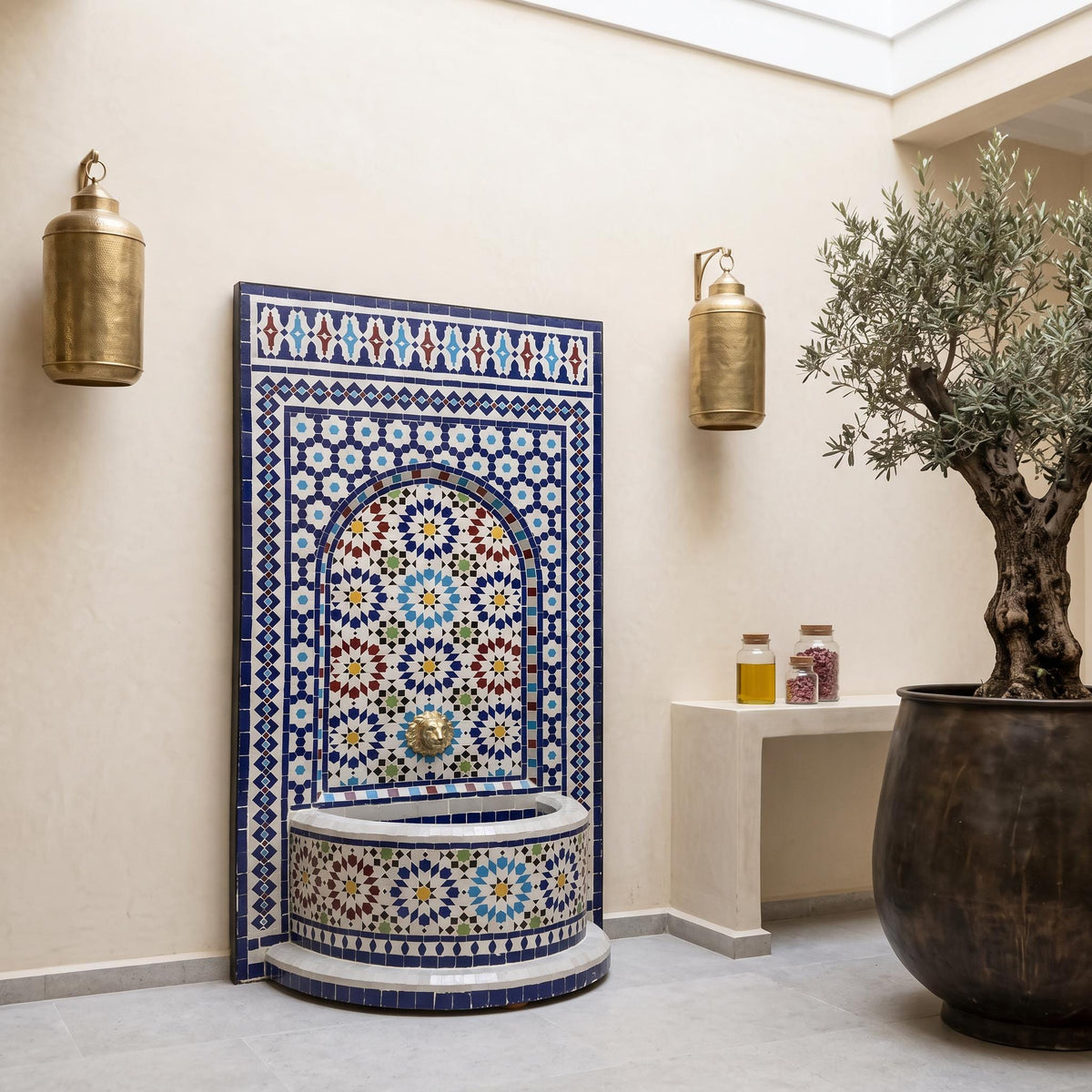 a decorative tile fountain with a mosaic design, flanked by two gold lanterns on the wall. The fountain is situated in a room with a white wall and a wooden floor. A potted plant is also present in the room, adding a touch of greenery to the space.