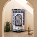 a decorative fountain with a mosaic design, surrounded by a green vase, a lantern, and a rug, all set against a beige wall with an arched doorway.
