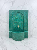 a green mosaic-tiled fountain with a gold-colored spout, set against a marble wall.