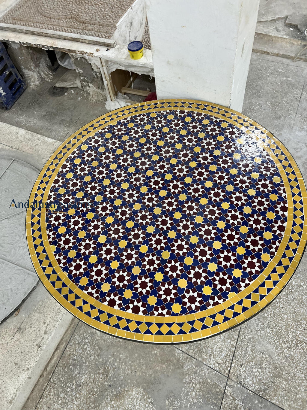 a circular table with a decorative tile pattern in shades of blue, yellow, and red. The table is situated on a concrete floor, and there is a white pillar visible in the background.