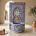 a decorative tile fountain with a blue and white mosaic design, situated in a room with a copper pot and a small potted plant.