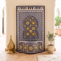 a decorative tile fountain with a blue and white mosaic design, situated in a hallway with a tiled floor. The fountain is surrounded by potted plants and a golden object, creating an elegant and visually appealing scene.