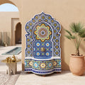 a decorative fountain with a mosaic design, situated in an outdoor setting with a table and potted plant nearby.