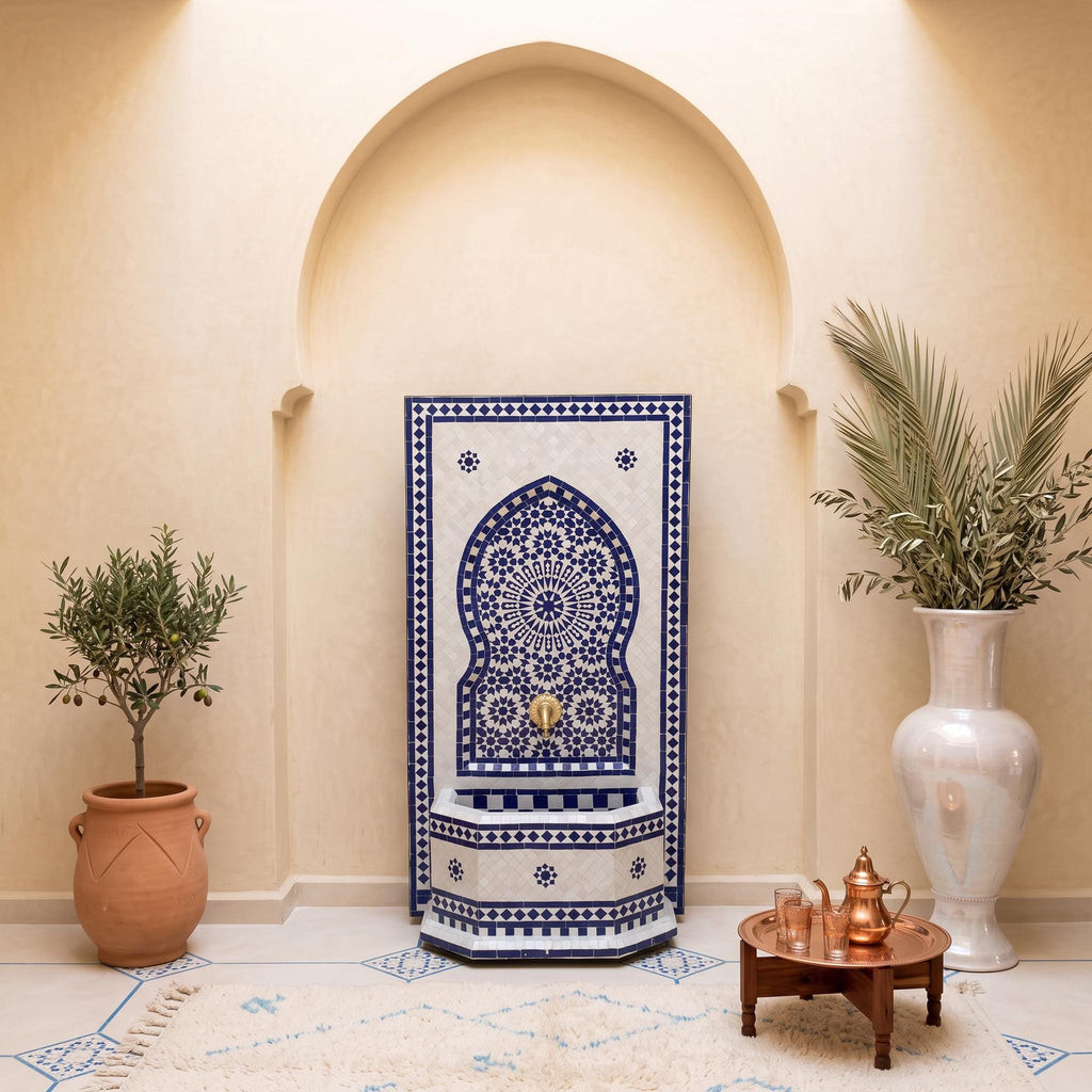 a room with a blue and white patterned tile wall, a white vase with a plant, a blue and white patterned tile fountain, and a wooden table with a copper kettle and tea set.