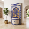 a decorative blue and white tile fountain with a gold-colored base, situated in a room with a potted plant and a wooden archway visible.