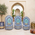 a courtyard with three ornate chairs adorned with blue and white mosaic patterns, situated in front of a doorway with arches. The chairs are surrounded by potted plants, and a small table is visible on the right side of the image.