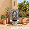 a decorative fountain with a mosaic design, surrounded by potted plants and a small tree. The fountain is situated in a courtyard with a tiled floor and a beige wall.