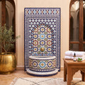 a decorative tile fountain with a blue and white mosaic design, situated in a room with a wooden table and a potted plant.