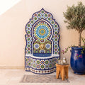 a colorful mosaic fountain with a blue and white pattern, situated in a room with a wooden stool and a potted plant.
