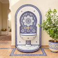 a decorative tile fountain with a blue and white pattern, situated in a room with a potted plant and a rug on the floor.
