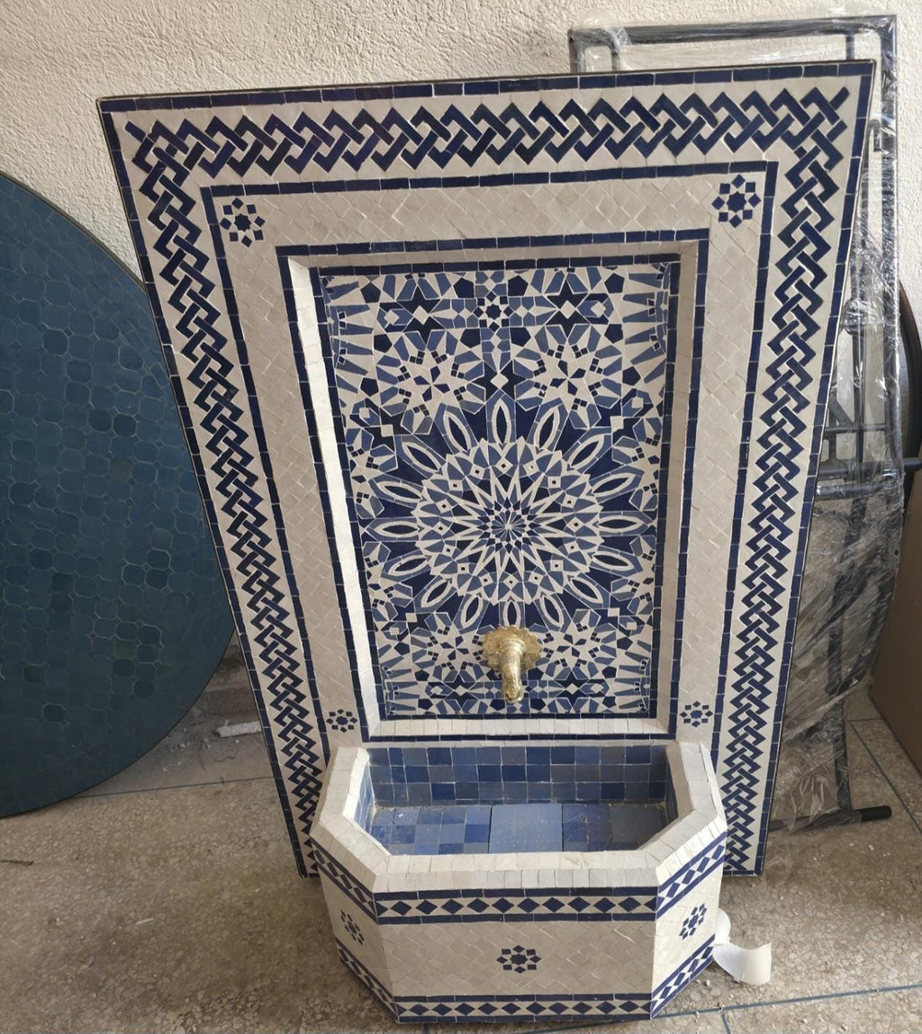Blue and white Moroccan mosaic fountain installed as a garden water feature on a patio, surrounded by ornamental grasses and succulents in terracotta wall pots.