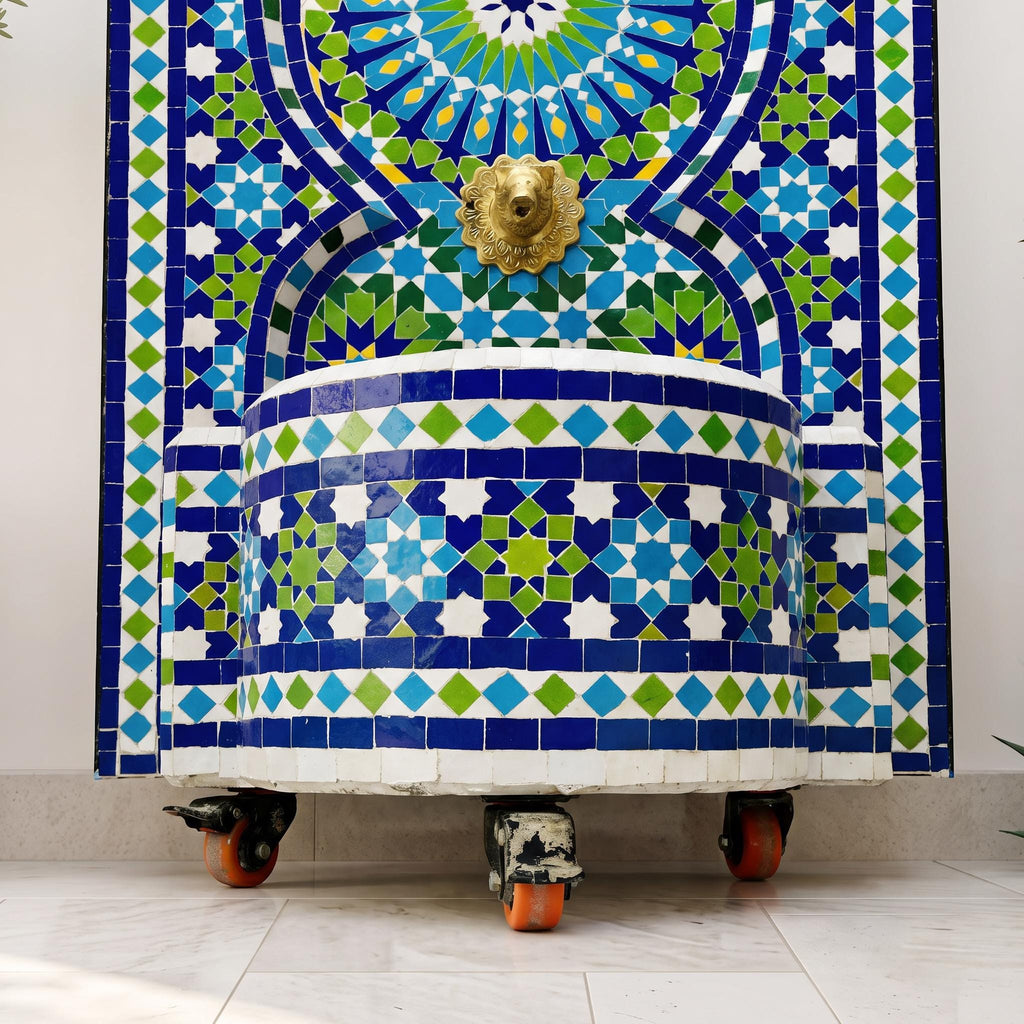 a decorative fountain with a mosaic tile design in shades of blue, green, and white. The fountain is mounted on a white pedestal with orange wheels, and a golden lion head is visible above the fountain.