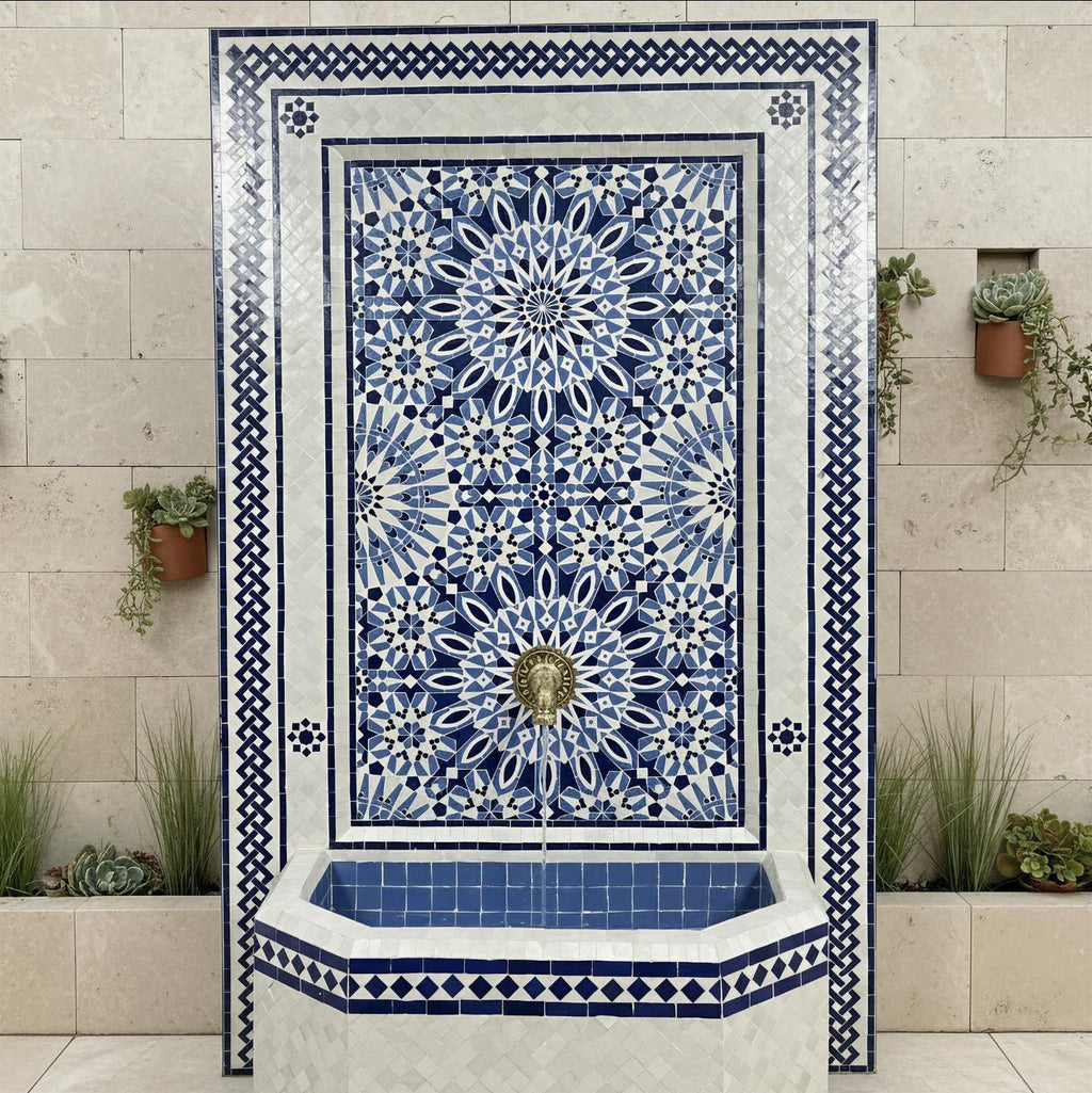 Handmade Moroccan mosaic wall fountain in blue and white zellige tile, front view, with a large geometric star pattern panel, brass lion head spout, and octagonal tiled basin against a stone wall.