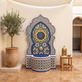 a decorative fountain with a mosaic design, surrounded by a potted plant and a small table with a bowl on it.