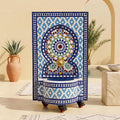 a decorative tile fountain with a blue and white geometric pattern, placed on a tiled floor. In the background, there is a potted plant and a bowl, creating a cozy and inviting atmosphere.