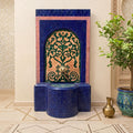 a blue and gold mosaic fountain with a decorative arch, situated in a room with a tiled floor and a potted plant.