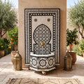 a decorative fountain with a black and white tile pattern, surrounded by potted plants and orange trees. The fountain is situated in a courtyard, with two lanterns placed on either side.