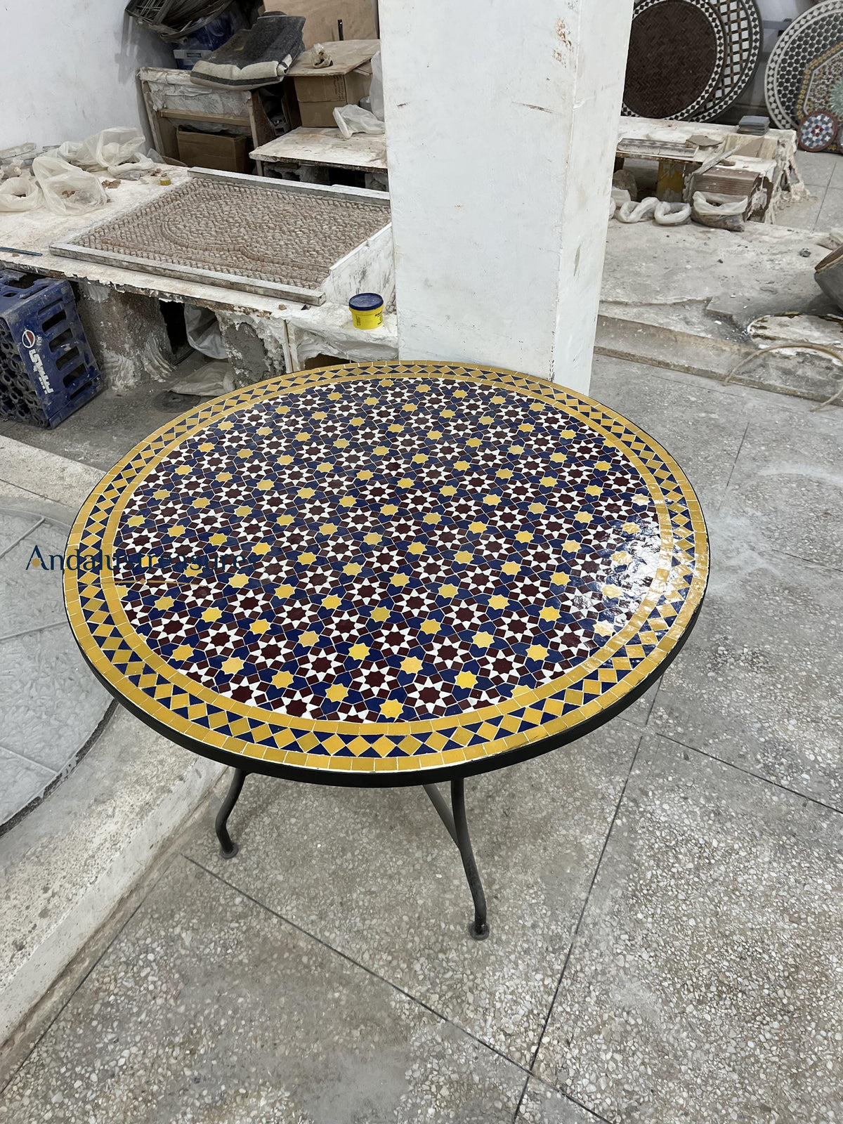 a round table with a decorative tile top, showcasing a vibrant pattern of blue and yellow colors. The table is situated in a workshop or studio setting, surrounded by various tools and materials.