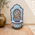 a colorful mosaic fountain with a blue and orange floral pattern, placed on a tiled floor next to a large gold vase.