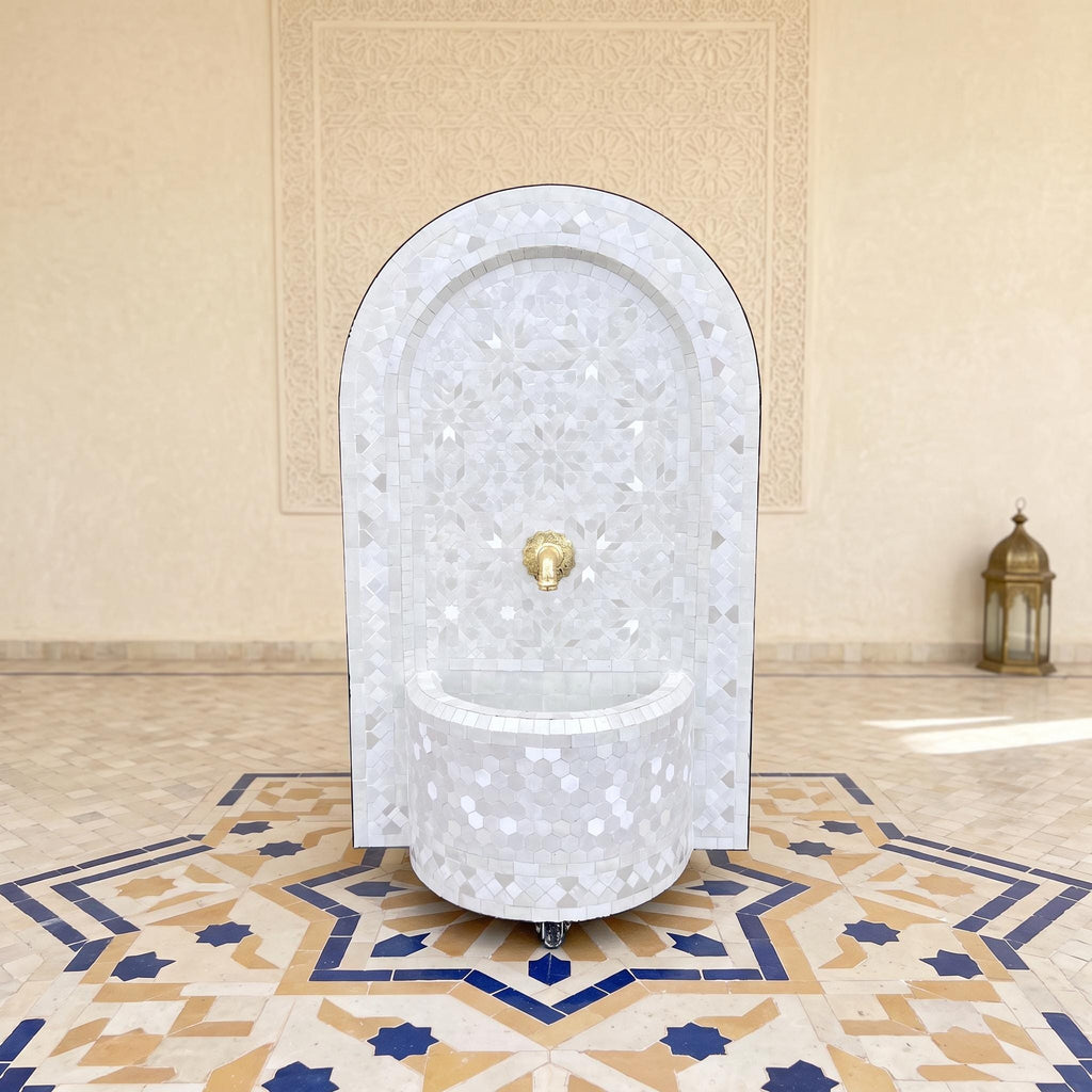 a white, ornate fountain with a gold-colored spout, situated in a room with a tiled floor and a lantern in the background.