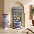 a decorative fountain with intricate blue and white tile patterns, situated in a room with a white archway. Next to the fountain is a large blue and white vase, and a potted plant is visible in the background.