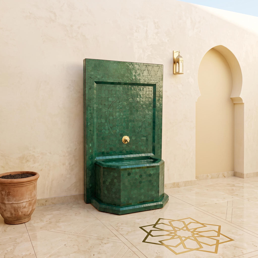 a green mosaic fountain with a gold-colored spout, situated in a courtyard with a beige wall and a geometric pattern on the floor.