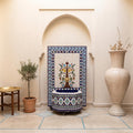 a decorative tile fountain with a blue and white border, situated in a room with a white archway. The room also includes a table with a plant and a large white vase.