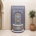 a decorative tile fountain with a blue and white mosaic design, situated in a room with a potted plant and a wall.