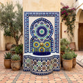 a decorative fountain with a mosaic design, surrounded by potted plants and a building with arched windows in the background.