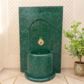a green mosaic tile fountain with a gold faucet, situated in a room with a potted plant and a geometric patterned wall.
