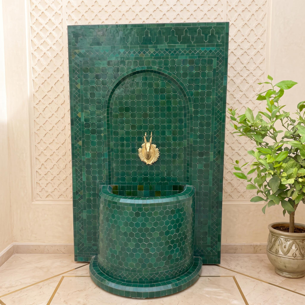 a green mosaic tile fountain with a gold faucet, situated in a room with a potted plant and a geometric patterned wall.