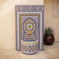 a decorative mosaic tile fountain with a blue and white border, situated in a room with a potted plant and a wall with a framed picture.