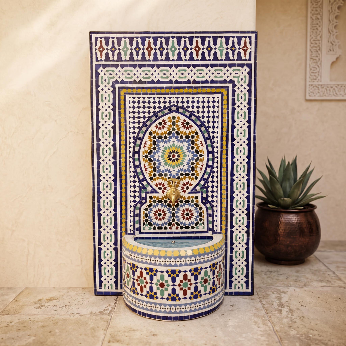 a decorative mosaic tile fountain with a blue and white border, situated in a room with a potted plant and a wall with a framed picture.