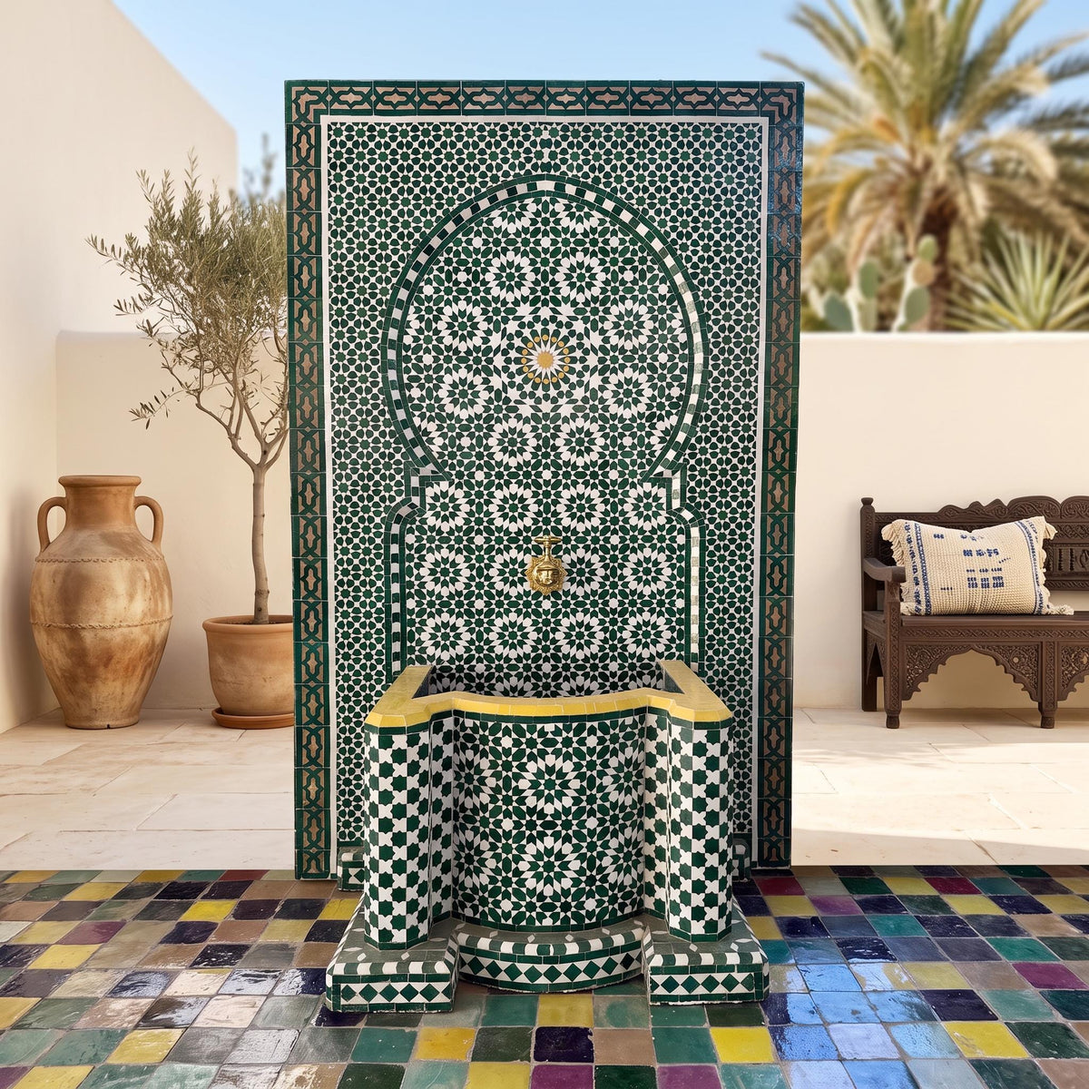 a decorative fountain with a mosaic tile design, situated in a courtyard with a bench and palm trees in the background.