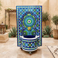a decorative fountain with a mosaic design in blue, green, and white colors, situated in a courtyard surrounded by potted plants and a rug.