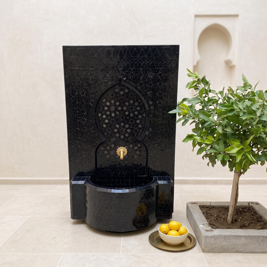 a black mosaic fountain with a gold faucet, situated next to a potted plant and a bowl of lemons on a tiled floor.