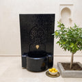 a black mosaic fountain with a gold faucet, situated next to a potted plant and a bowl of lemons on a tiled floor.