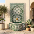a decorative fountain with a green and white tile pattern, surrounded by potted plants and a building with arched windows.