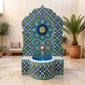 a decorative fountain with a mosaic design in blue, green, and yellow colors, situated in a room with a beige wall and potted plants.