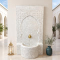 a white mosaic fountain with a gold faucet, situated in a courtyard with arched doorways and potted plants.