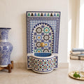 a decorative tile fountain with a blue and white pattern, placed on a white tiled floor. Next to the fountain is a blue and white vase, and a stack of towels is visible in the background.