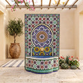 a colorful mosaic fountain with a circular design and a sunburst motif, surrounded by potted plants and a wooden pergola.