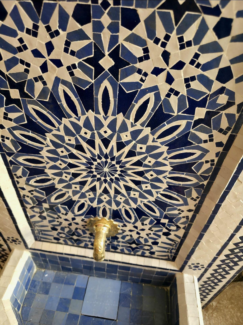 Angled side view of the blue zellige Moroccan fountain showing the raised mosaic panel, wall mounted design, and octagonal water basin, set on a travertine patio with potted succulents.