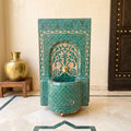 a decorative green tile fountain with a gold-colored vase on a wooden stand, placed on a marble floor.