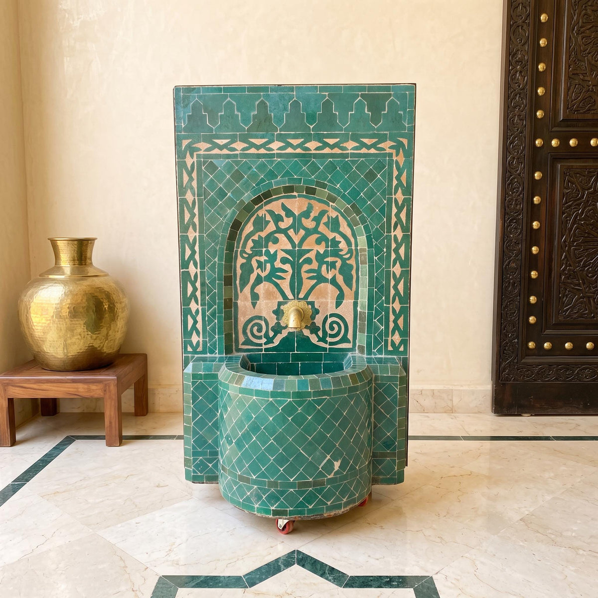 a decorative green tile fountain with a gold-colored vase on a wooden stand, placed on a marble floor.