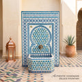 a beautifully crafted, authentic handmade Zellice fountain, with a blue and white tile pattern and a copper lantern on the left side. The fountain is situated in a courtyard with a tiled floor and a potted plant nearby.