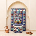 a decorative fountain with a mosaic design, situated in an arched doorway. Next to the fountain is a copper pitcher, and a pair of brown shoes are placed on the floor in front of the fountain.
